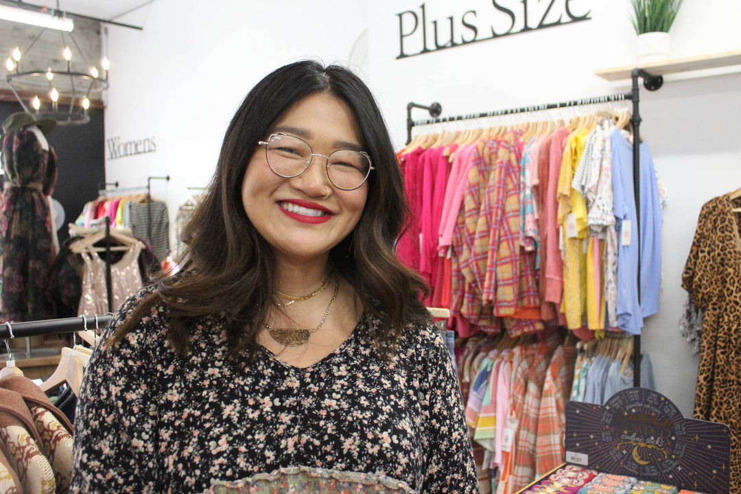 Asian woman business owner emily brooker of esme and elodie standing in front of racks of plus sized clothing in her renton boutique