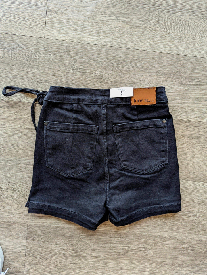 back of judy blue black skort showing the pockets and the side tie laid on floor